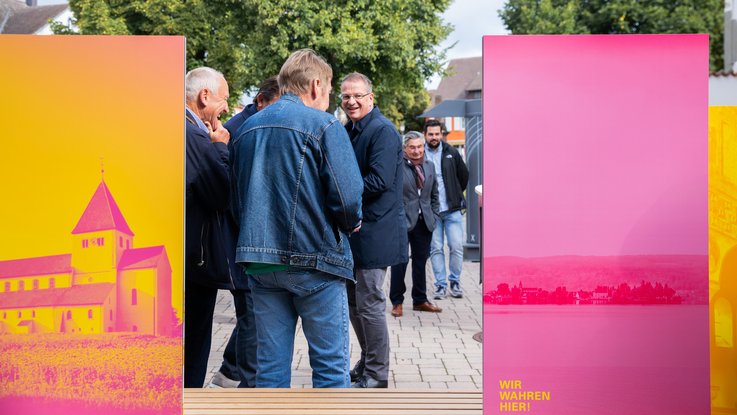 VISUELL Szenografie: Wahre Wunderbänke: Eine Gruppe von Besuchenden steht vor einer Sitzbank, die Rückseite zweier Informationstafeln mit Bildmotiven in magenta und gelb ist sichtbar