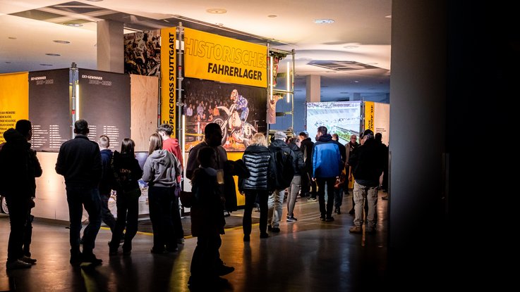 VISUELL Szenografie: Ausstellung: Zahlreiche Besucherinnen und Besucher flanieren durch die offene Szenografie; großflächige Banner in Gelb und Schwarz strukturieren den Raum.