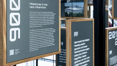 VISUELL Szenografie: Jubiläumsausstellung: Detail einer Informationstafel mit weißer Typografie auf anthrazitfarbenem Grund in einem Holzrahmen, im Hintergrund sind weitere Informationstafeln zu sehen.