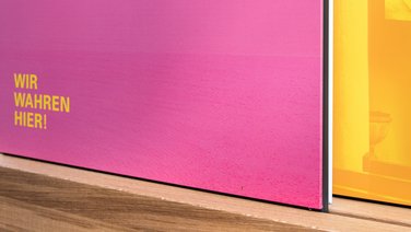 VISUELL Szenografie: Wahre Wunderbänke: Detailaufnahme der Rückseite zweier Informationstafeln mit Bildmotiv in magenta und gelb mit der Aufschrift WIR WAHREN HIER!